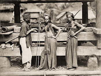 Tri batakaj militistoj kun lancoj kaj spadoj antaŭ ligna konstruaĵo en Indonezio (ĉ. 1870)